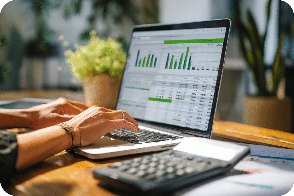 Uma mulher trabalhando em um laptop sobre uma mesa de madeira, digitando enquanto visualiza gráficos financeiros e tabelas de métricas de negócios. Uma calculadora está visível em primeiro plano.