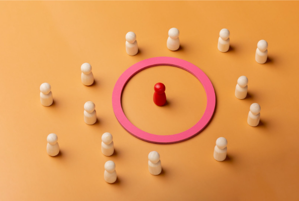 Imagem conceitual de liderança e destaque. Um peão de madeira vermelho está posicionado ao centro de um círculo rosa, cercado por diversos peões de madeira clara dispostos em círculo sobre um fundo laranja. Representação visual de protagonismo, individualidade e estratégia de marca.