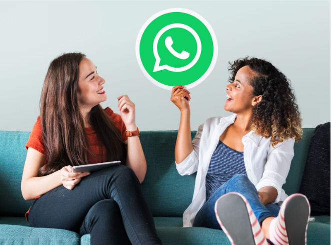 Duas mulheres sorridentes sentadas em um sofá azul conversando. Uma delas segura um círculo com o logotipo do WhatsApp e a outra segura um tablet. A imagem representa o uso do WhatsApp Business para vendas, engajamento de clientes e comunicação digital moderna