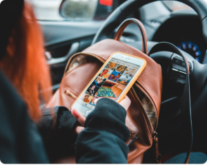 Pessoa sentada no banco de um carro segurando um smartphone que exibe um feed de produtos no Instagram. O celular está posicionado em frente a uma bolsa de couro marrom, simulando uma jornada de compra mobile em um ambiente cotidiano.