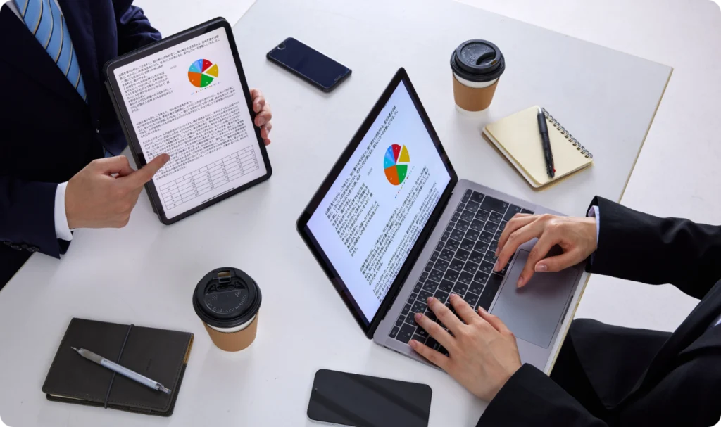Visão superior de uma reunião de negócios focada em análise de dados. Uma pessoa utiliza um tablet para apresentar gráficos de pizza coloridos, enquanto outra trabalha em um notebook visualizando as mesmas métricas. Sobre a mesa branca, há xícaras de café, cadernos de notas e smartphones, simbolizando uma operação híbrida eficiente e a tomada de decisão baseada em Business Intelligence