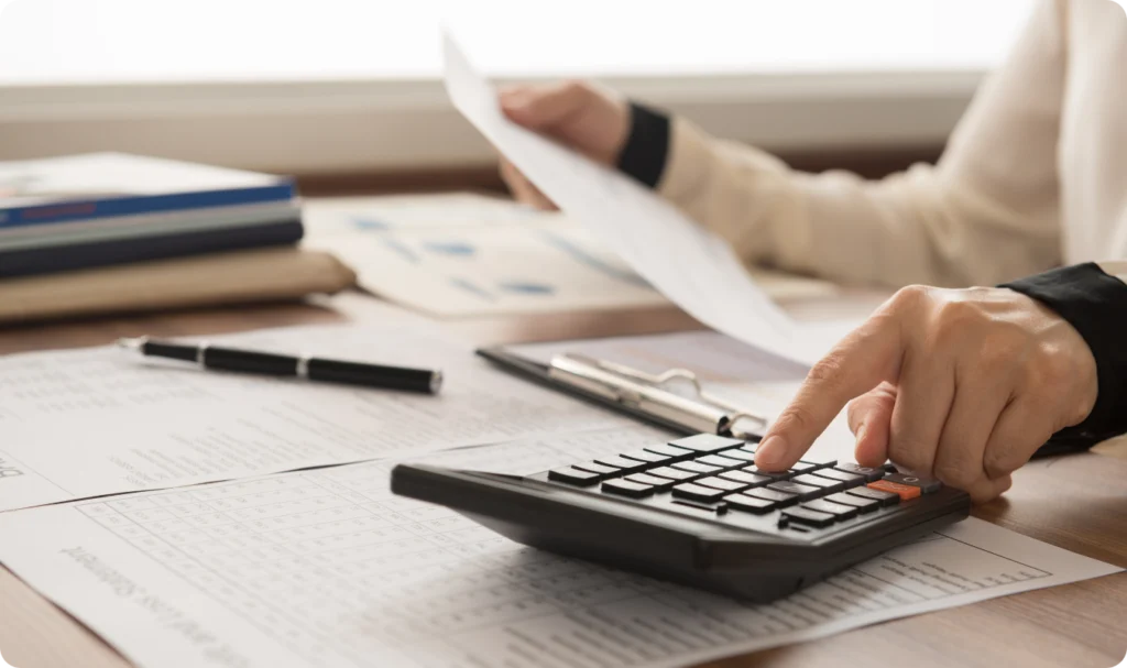 Close-up das mãos de um profissional de contabilidade utilizando uma calculadora financeira sobre uma mesa repleta de planilhas e relatórios de demonstrativos de resultados. A imagem ilustra a análise de eficiência financeira, o cálculo preciso de margens de lucro e o planejamento estratégico para o crescimento sustentável de uma empresa.
