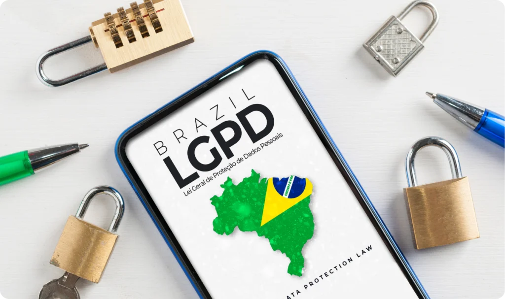 Visão superior de um smartphone sobre uma mesa branca, exibindo na tela o texto 'BRAZIL LGPD - Lei Geral de Proteção de Dados Pessoais' junto a um mapa do Brasil com as cores da bandeira. Ao redor do aparelho, há diversos cadeados metálicos de diferentes tamanhos, abertos e fechados, e canetas, simbolizando a segurança jurídica, a privacidade do consumidor digital e a implementação de protocolos de proteção de dados.