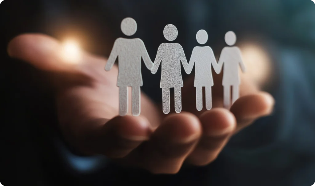 Uma mão aberta em concha sustenta quatro figuras humanas de papel ou acrílico que estão de mãos dadas. Ao fundo, luzes suaves (bokeh) sugerem um ambiente profissional e acolhedor. A imagem simboliza a retenção de talentos, a união da equipe e o cuidado da gestão com o capital humano dentro de uma organização